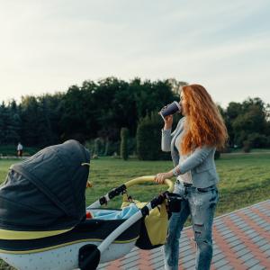 A young mother enjoying the park walk in the morning, drinking a hot tea, with her little child relaxing in his baby trolley.