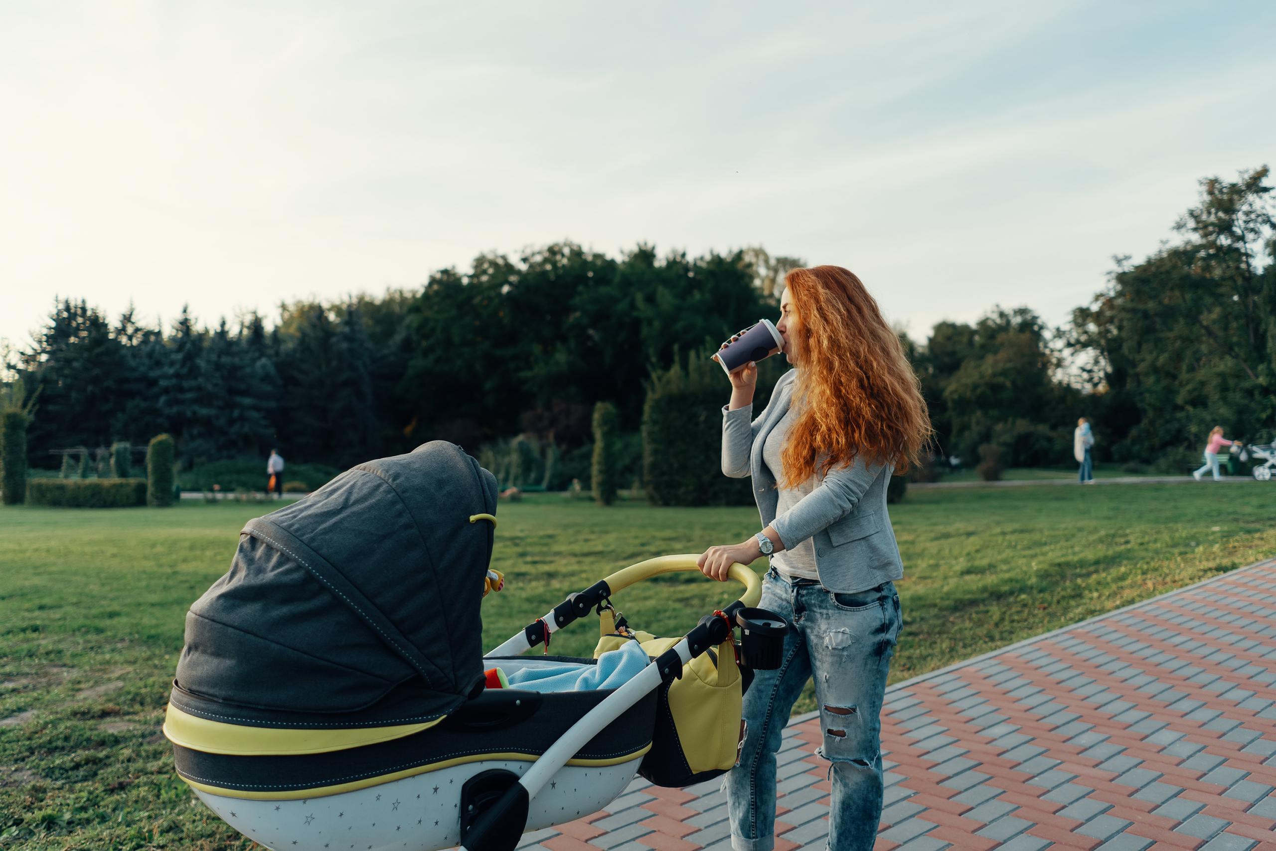 A young mother enjoying the park walk in the morning, drinking a hot tea, with her little child relaxing in his baby trolley.