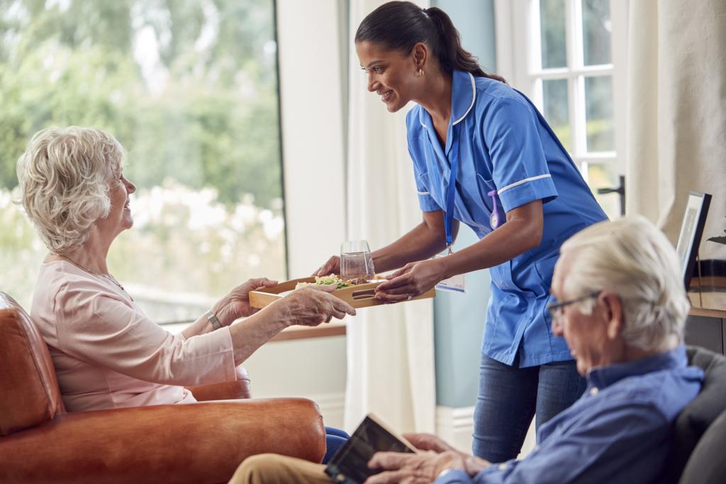 Female Care Worker In Uniform Bringing Meal On Tray To Senior Woman Sitting In Lounge At Care Home