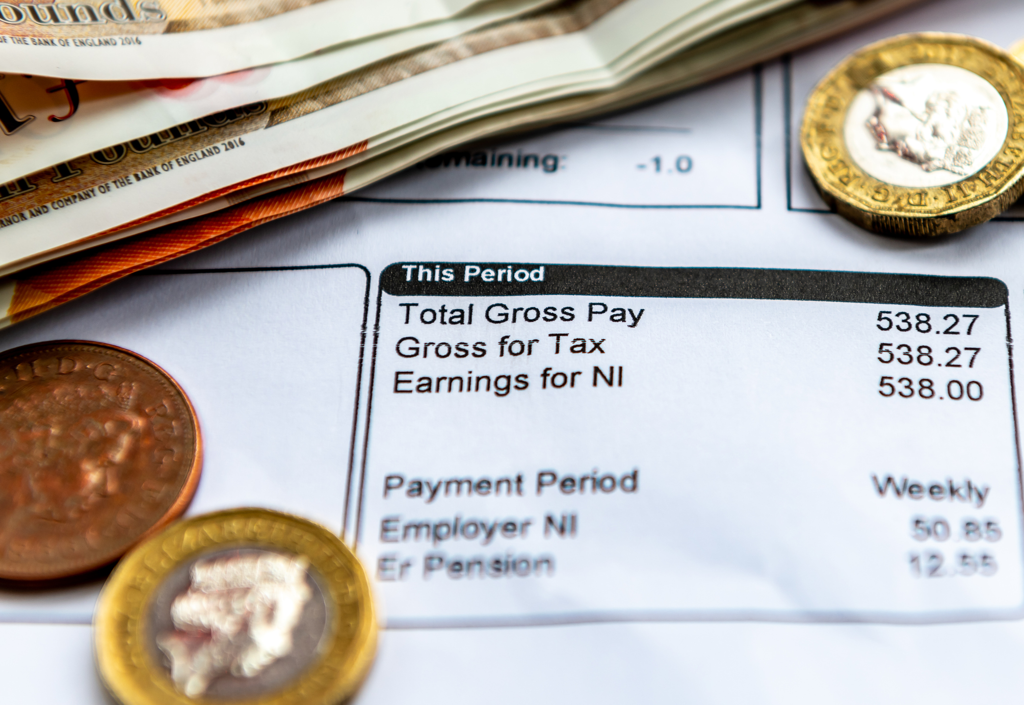 A payslip showing pay as you earn tax deductions, national insurance and pension contributions with some bank notes and coins.